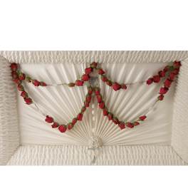 Garland of red rosebuds draped inside a white pleated casket lid with a hanging rosary cross.