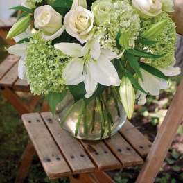 White lilies, pale roses, and green hydrangeas arranged in a clear glass vase on a wooden chair outdoors