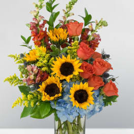 Sunflowers, orange roses, and blue hydrangeas in a clear glass vase
