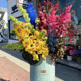 Tall colorful floral arrangement in a cylindrical box with orchids and feathery accents