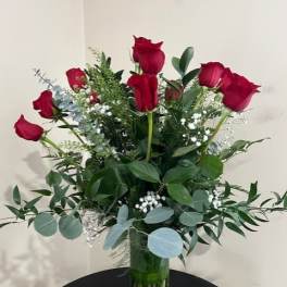 Tall arrangement of long-stem red roses with white filler flowers in a clear glass vase