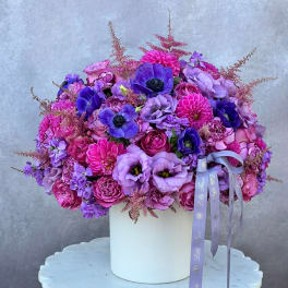 Purple and pink floral arrangement in a white box with ribbon tails