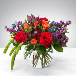 Low glass bowl of red roses, orange blooms, and purple flowers with trailing green accents