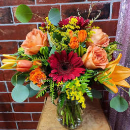 Mixed bouquet of roses, gerbera daisies, lilies, and yellow filler in a glass vase