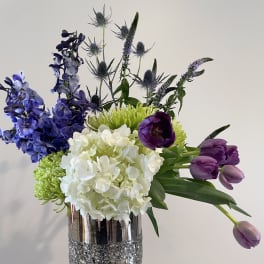 Mixed bouquet with purple tulips, blue delphinium, and white hydrangea in a silver vase