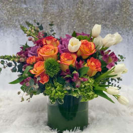 Low cylinder vase of orange roses, purple blooms, white tulips and green accents