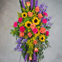 Tall standing spray of sunflowers, orange roses, pink carnations, and purple blooms on an easel.