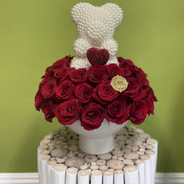 Red roses arranged around a pearl-covered teddy bear in a white vase