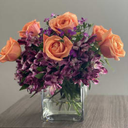 Peach roses and purple flowers arranged in a square glass vase