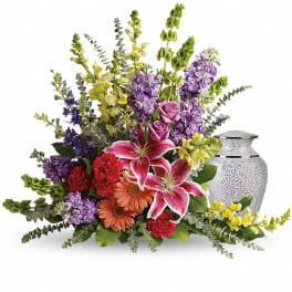 Colorful floral arrangement with lilies, roses, and gerbera daisies beside a decorative urn