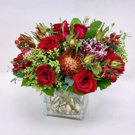 Low arrangement of red roses and protea blooms in a clear glass cube vase with berries