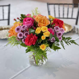 Bright mixed arrangement of roses, carnations, and daisies in a clear glass vase on a table.