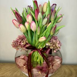 Pink and purple tulips in a round glass vase with a burgundy ribbon