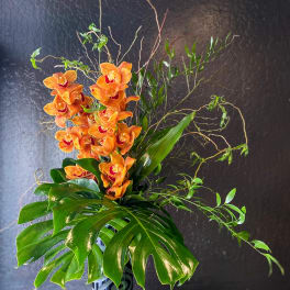 Tall arrangement of orange orchids with large monstera leaves in a patterned black cylinder vase