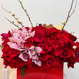 Red roses with pink orchids in a red box, accented by flowering branches.