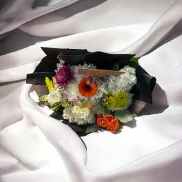 Mixed bouquet with white hydrangeas, orange gerbera daisies, and purple blooms in black wrap