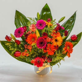 Bright mixed bouquet of orange gerbera daisies, red roses, and pink blooms in a glass vase with ribbon.