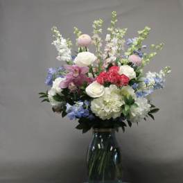 Tall mixed arrangement of white hydrangeas, roses, pink orchids, and blue delphinium in a clear glass vase