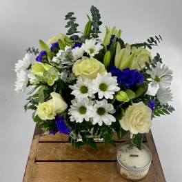 White and cream bouquet with purple accents in a glass vase beside a white tin candle