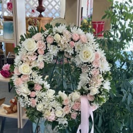 Heart-shaped wreath of white and blush flowers on a stand with a pink ribbon
