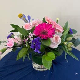 Low glass vase of pink gerbera daisy, roses, and mixed blooms with purple and yellow accents.