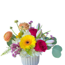 Cheerful mixed arrangement with yellow gerbera, pink roses, ranunculus and tulips in a white ceramic vase.