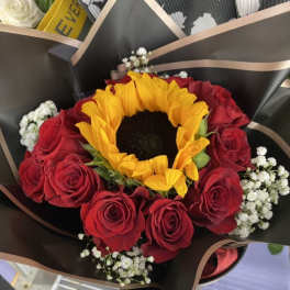 Hand-tied bouquet with a large yellow sunflower, red roses, and white filler flowers in black wrapping paper