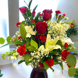 Red roses, white lilies, and red carnations arranged with filler blooms in a red glass vase.