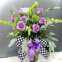 Purple roses in a glass vase with black-and-white checkered ribbon and a purple bow