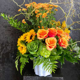 Tall orange and yellow arrangement with roses, sunflowers and mums in a white vase with curly branches