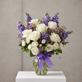 White roses and purple flowers arranged in a glass vase with a purple ribbon