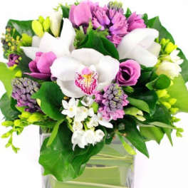 Compact bouquet of white orchids and purple flowers in a square glass vase lined with broad green leaves