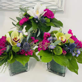 Three floral arrangements in square glass vases with orchids and bright pink blooms