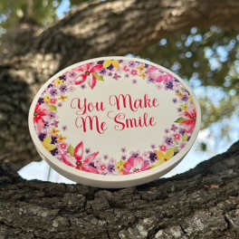 Oval floral sign reading "You Make Me Smile" on a tree branch