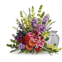 Mixed bouquet with lilies, roses, gerbera daisies, and purple flowers beside a silver urn