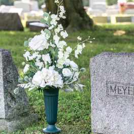 White floral arrangement in a dark green cemetery vase beside gravestones