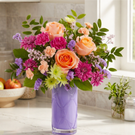 Mixed bouquet of peach roses, pink blooms, and purple flowers in a lavender vase