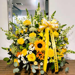 Tall yellow and white sympathy arrangement with roses, sunflowers, and snapdragons in a basket with memorial ribbon.