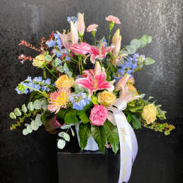 Pastel arrangement of pink lilies, roses, carnations, and blue flowers in a white vase with a white ribbon.