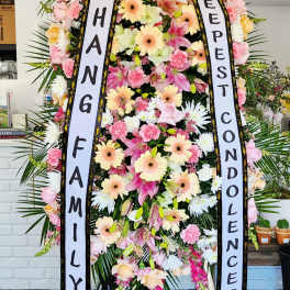 Tall standing spray of pink lilies, carnations, and peach gerberas with condolence ribbons on an easel.