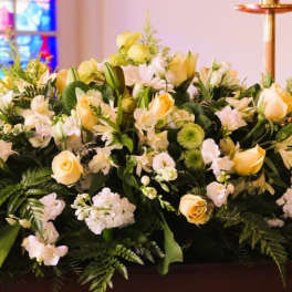 Long arrangement of white and pale yellow roses, lilies, and daisies on a casket in a church setting