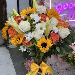 Bouquet of sunflowers, roses, lilies, and white daisies in a glass vase with a yellow bow