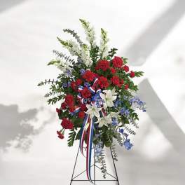 Standing floral spray with red carnations, white lilies, and blue accents on a metal easel