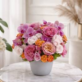Low round arrangement of pink roses, orchids, and orange spray roses in a white pot on a marble table