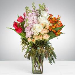 Tall mixed bouquet of white and peach roses, pink tulips, and orange alstroemeria in a clear glass vase