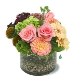 Short arrangement of pink roses, dahlias, and yellow gerberas in a glass vase with pebbles.