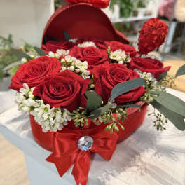 Red roses arranged in a heart-shaped box with white filler flowers and ribbon bows