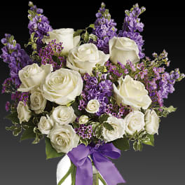 White roses and purple flowers arranged in a glass vase with a purple ribbon