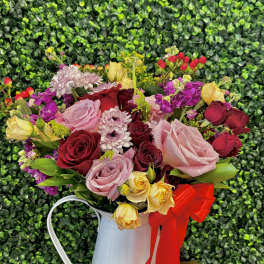 Mixed bouquet of roses and small blooms in a white metal pitcher with a red ribbon