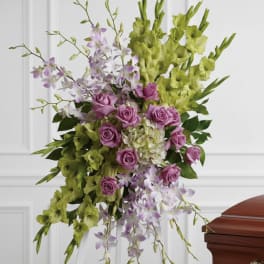 Standing spray of green gladiolus, lavender roses, and white orchids on a white easel beside a casket.